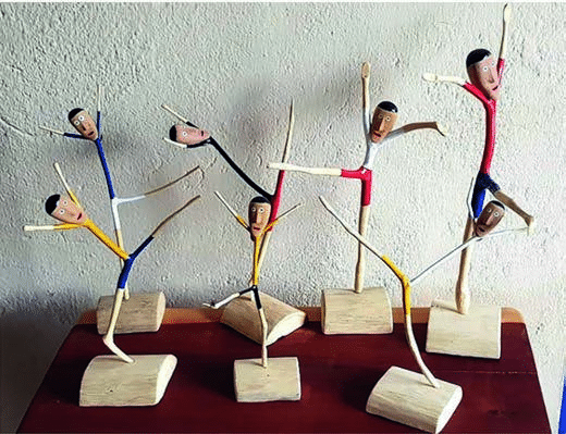 Fotografia de esculturas feitas com gravetos de madeira, coloridas e montadas em bases, representando figuras humanas em diferentes posições de dança.