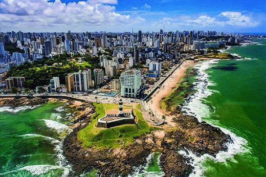 Fotografia aérea da orla de Salvador, com grande concentração de edifícios altos junto à faixa litorânea e ao Farol da Barra.