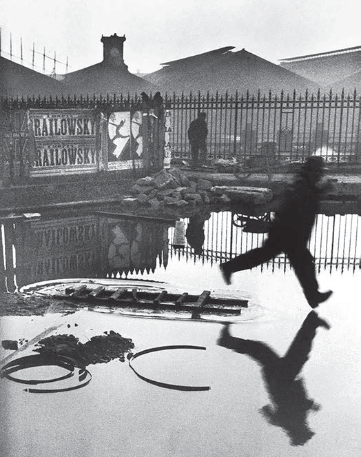 Fotografia em preto e branco de uma pessoa saltando sobre água junto à estação de Saint-Lazare. Sua silhueta e as grades se refletem na água; um cartaz com uma figura em movimento e formas curvas do chão participam da composição.