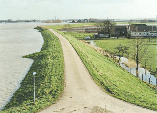 Fotografia de uma estrada sobre um dique coberto de grama. À esquerda há um grande corpo de água; à direita aparecem canal, casas e áreas de terreno mais baixo.